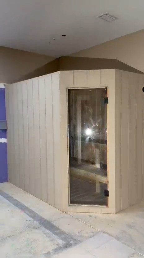 Indoor sauna in tan vertical grooved siding with an angled front corner and a full-height glass door, in an unfinished room