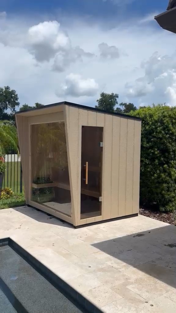 Beige vertical-groove outdoor sauna with a tinted glass door and a glass end wall on a travertine patio by a pool