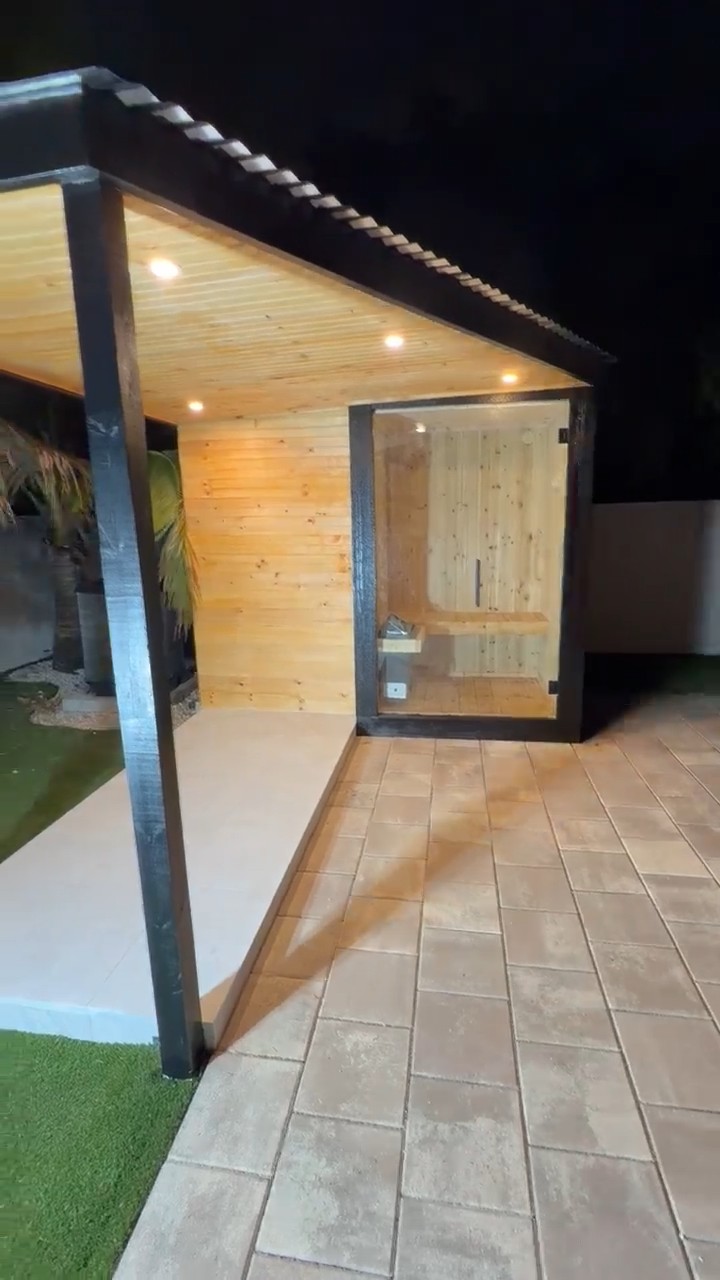 Black outdoor sauna at night with a covered porch, wood-lined ceiling with downlights and a glass-front sauna room