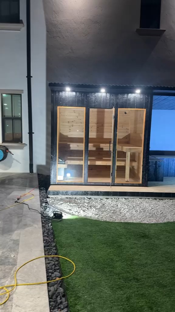Black vertical-board outdoor sauna at night, its three tall glass panels lit warm from inside, puck downlights above