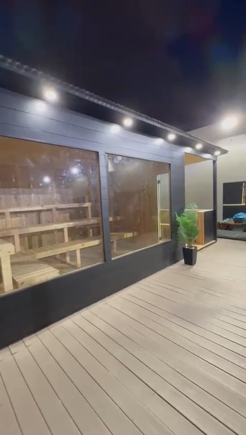 Black-sided sauna at night with two picture windows, soffit downlights and a covered porch at one end on a grey deck