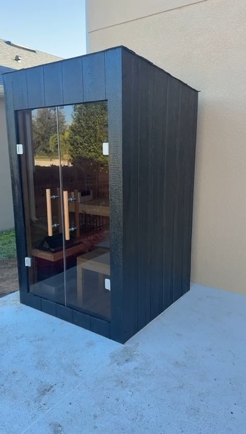 Compact sauna clad in black vertical boards with a pair of full-height glass doors, on a concrete pad by a stucco wall