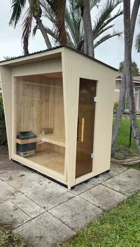 Compact sauna with cream siding, dark corrugated shed roof, glass front panel and glass side door on a paver pad