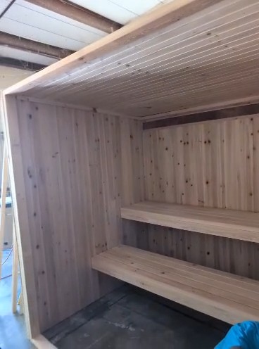 Open-front sauna room mid-build with knotty vertical wall boards, a board ceiling and two-tier slatted benches