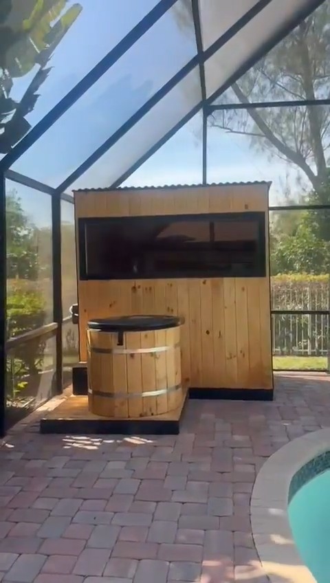 Cabin sauna with vertical knotty boards and a long dark window, wooden plunge tub on a deck, in a screened pool enclosure