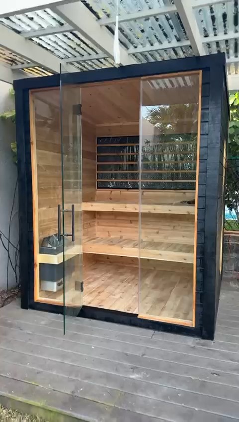 Black-clad cube sauna with full-height glass front and open door under a white pergola, showing two-tier benches inside