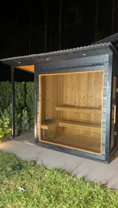 Black-clad sauna at night with a wide glass front showing two benches, beside a covered alcove under a corrugated roof