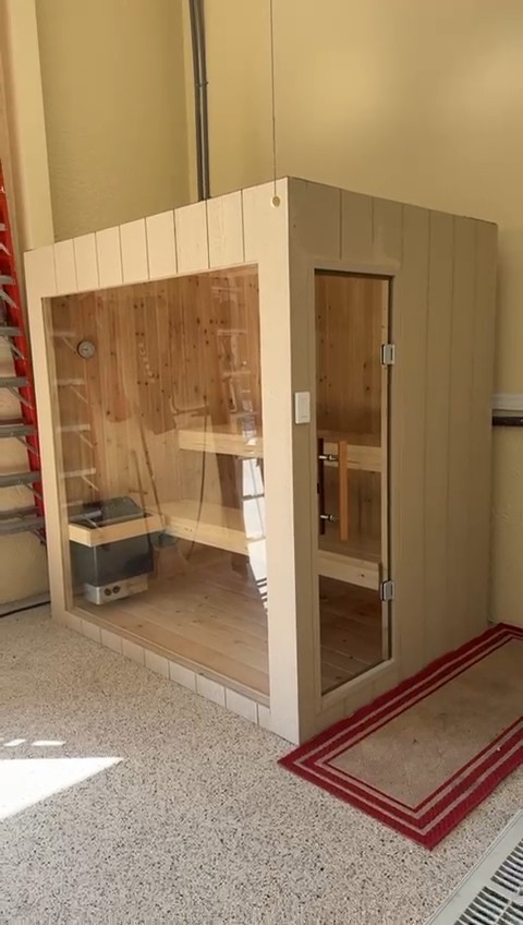 Freestanding indoor sauna in a garage with a greige vertical-board shell, large glass front and frameless glass door