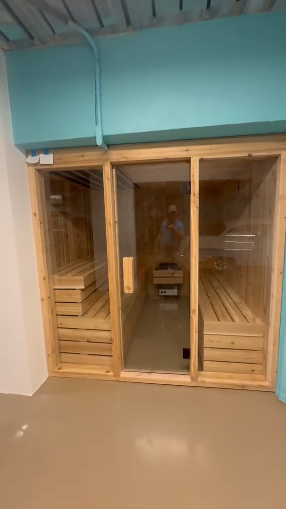 Indoor sauna with a three-panel glass front in a light wood frame under a teal soffit, benches and heater visible inside