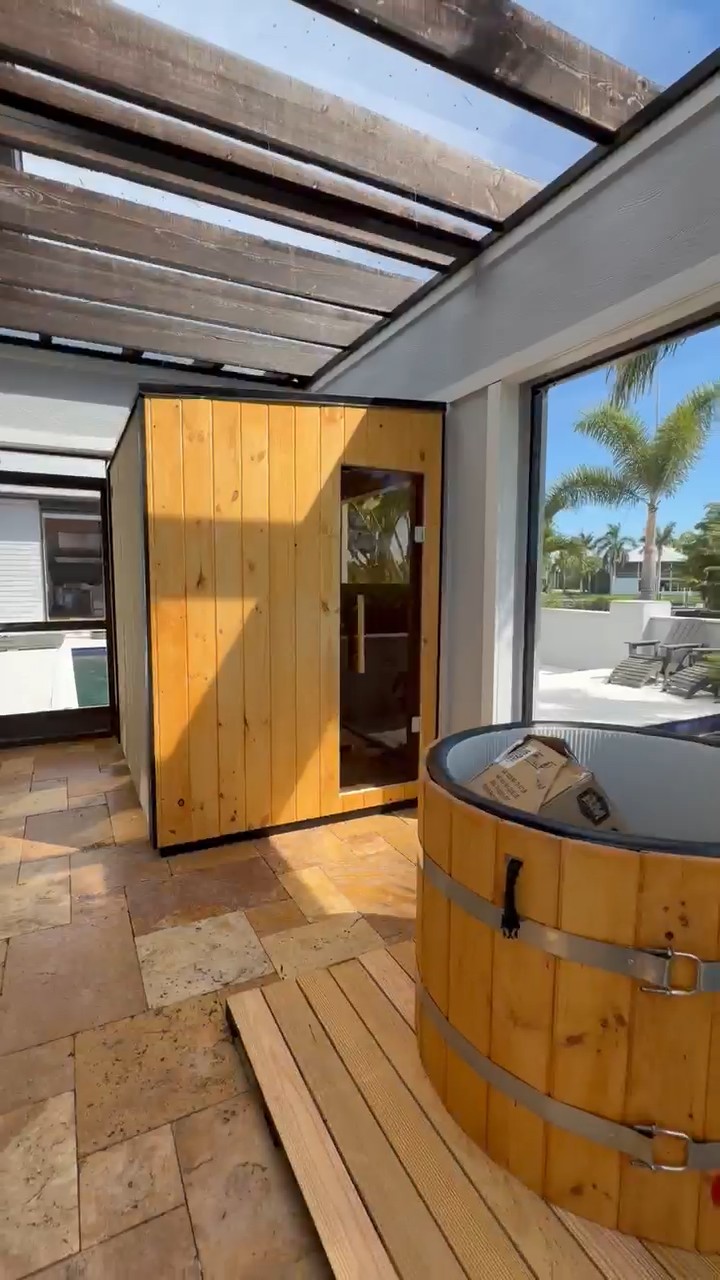 Cabinet sauna with vertical wood boards and a tinted glass door in a glass-roofed lanai, cold plunge in the foreground
