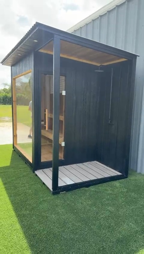 Black-clad sauna under a corrugated metal roof, with a glass door onto an open shower bay, rainfall shower and slatted deck