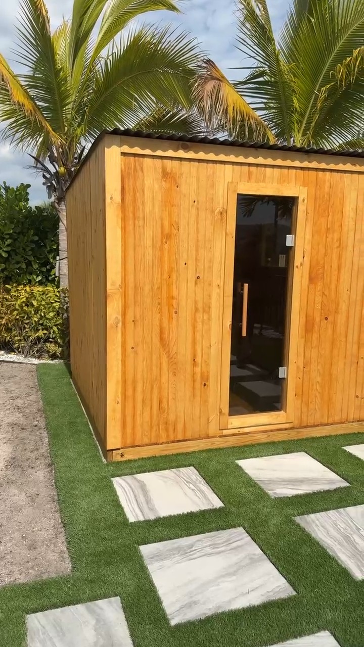 Honey-stained outdoor sauna with a full-height glass door and black corrugated roof on turf with white pavers and palms