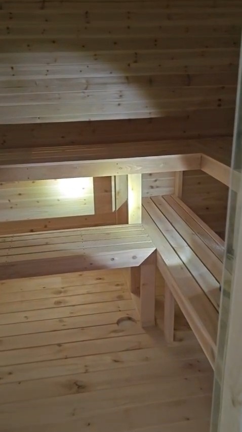 Inside the sauna: two-tier L-shaped slatted benches, light glowing under the upper bench, pale knotty horizontal wall boards