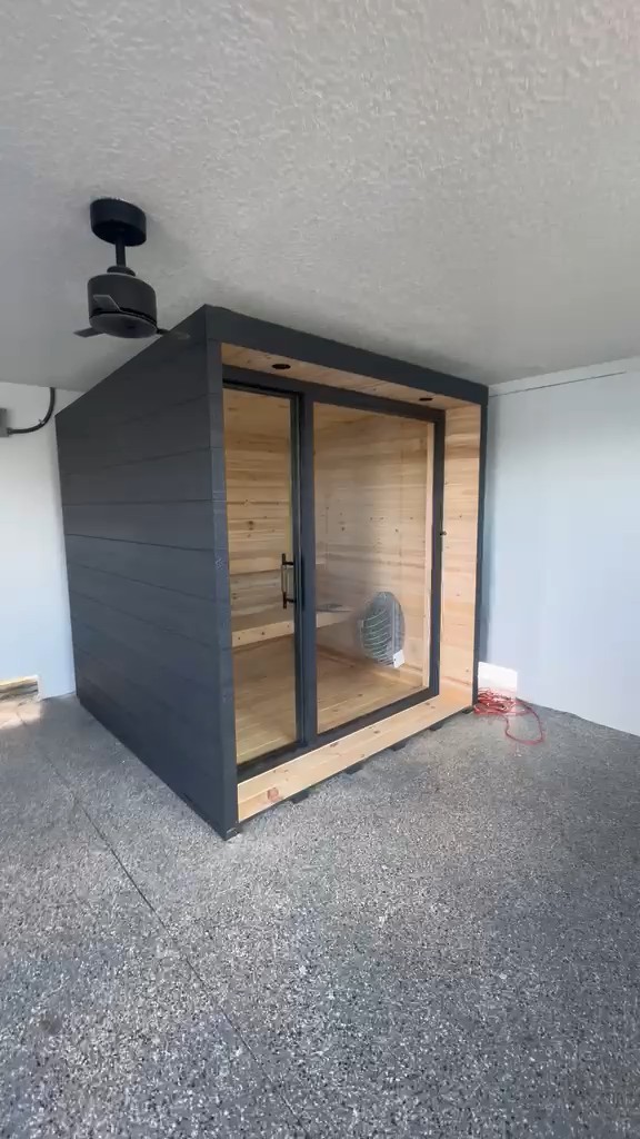 Indoor sauna seen from the front left: matte-black board side and a black-framed glass front under a natural-wood header