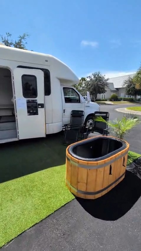 White shuttle bus mobile sauna with side door open, oval wooden cold plunge tub and potted palm on turf beside it