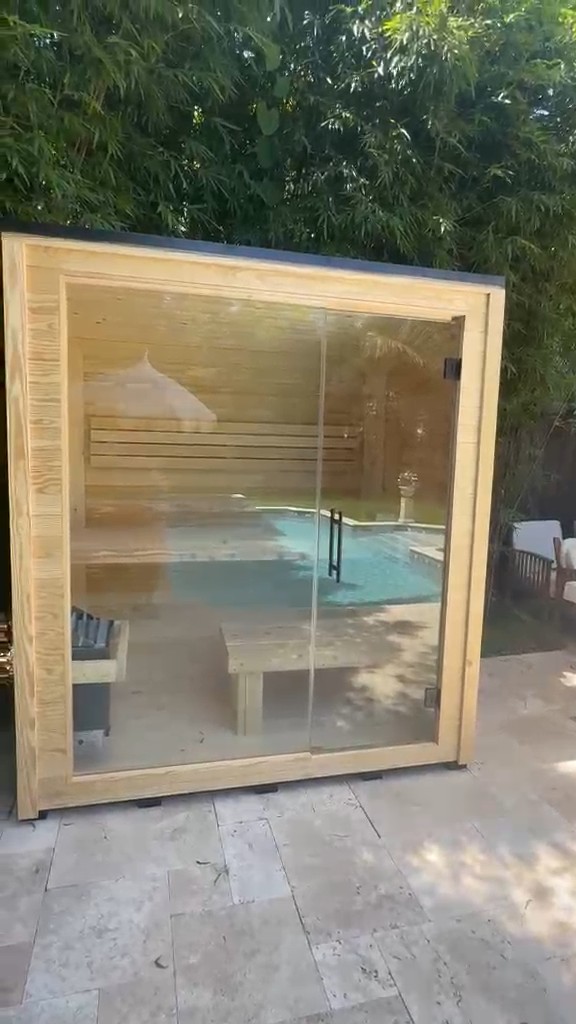 Square outdoor sauna with a pale wood frame, full glass front and frameless door on a travertine patio by a bamboo hedge