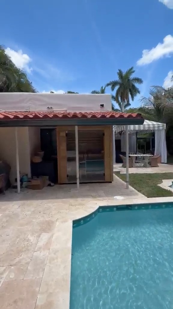 Box sauna with a glass front under a terracotta-tiled patio roof, seen across a pool and travertine deck