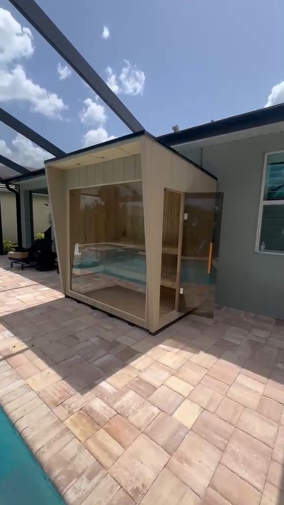 Cream shed-roof sauna with a large picture window and open tinted glass door on a paver pool deck under a screen cage