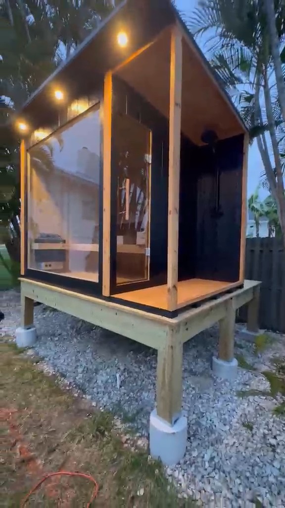 Black-clad sauna with full-height glass front and open shower bay on a raised wood deck at dusk