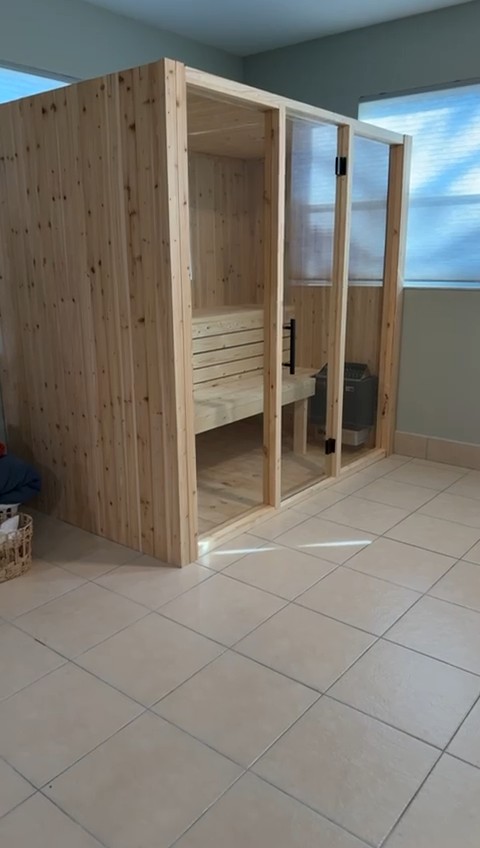 Freestanding knotty wood indoor sauna with a frameless glass door and fixed glass panel on a tiled floor