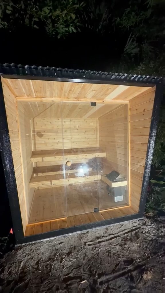 Outdoor sauna at night: glass front set back in a thick black frame, ribbed roof edge, lit pale-wood benches inside