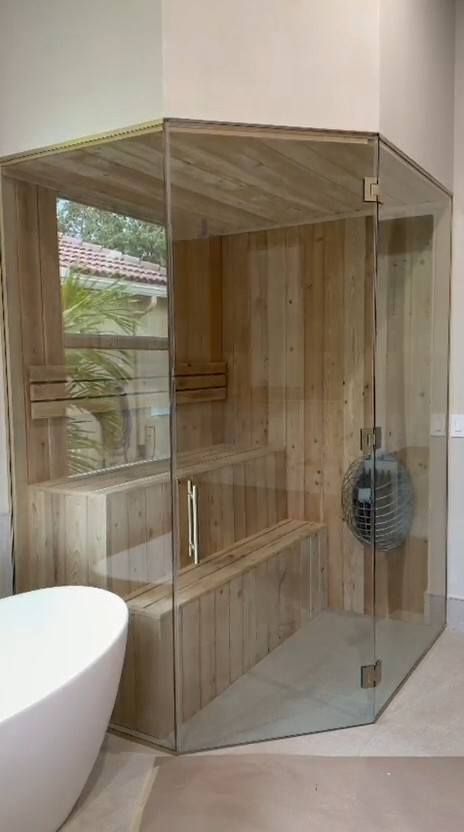 Corner sauna behind a three-panel frameless glass front, next to a white freestanding tub in a bathroom