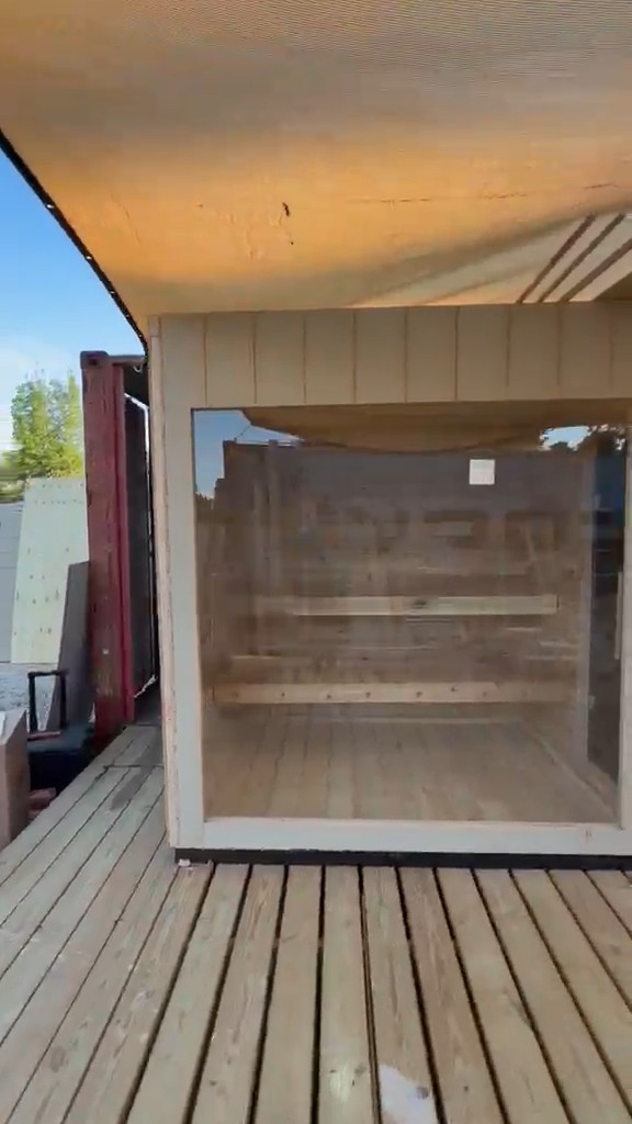 Sauna front: one large glass pane in a sand-toned frame, two benches visible inside, on a deck under a shade sail