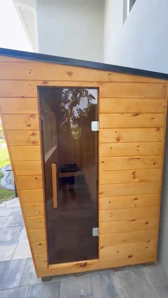 Compact outdoor sauna clad in horizontal knotty boards, with a full-height bronze-tinted glass door, set on grey pavers