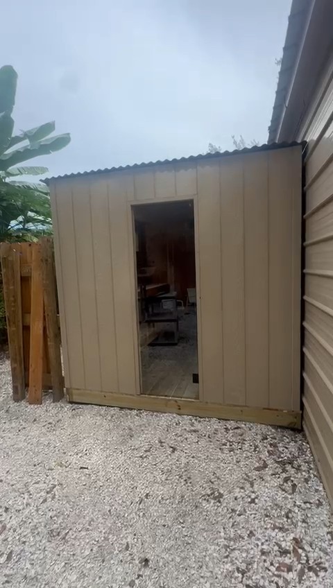 Tan vertical-panel sauna cabin with a corrugated metal roof and full-height glass door on a gravel side yard