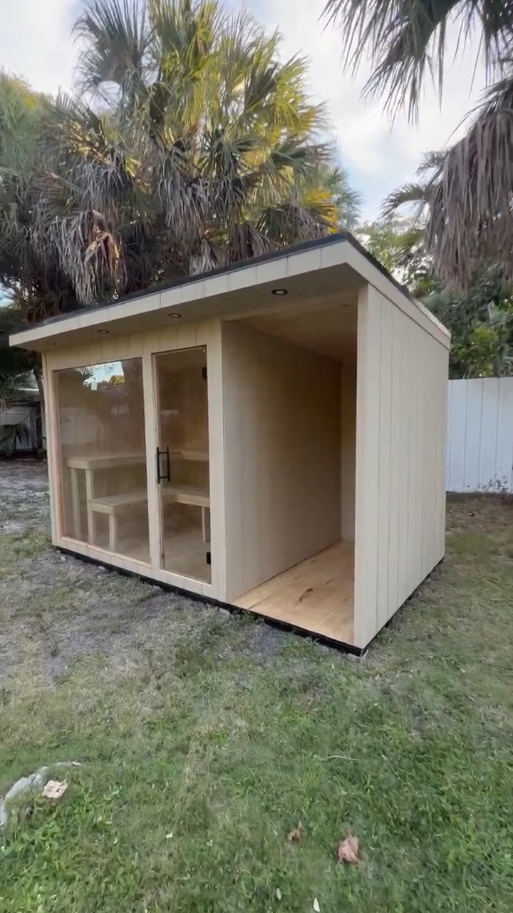 Glass-front sauna and open shower bay under a single-slope roof, three-quarter view on a backyard lawn with palms