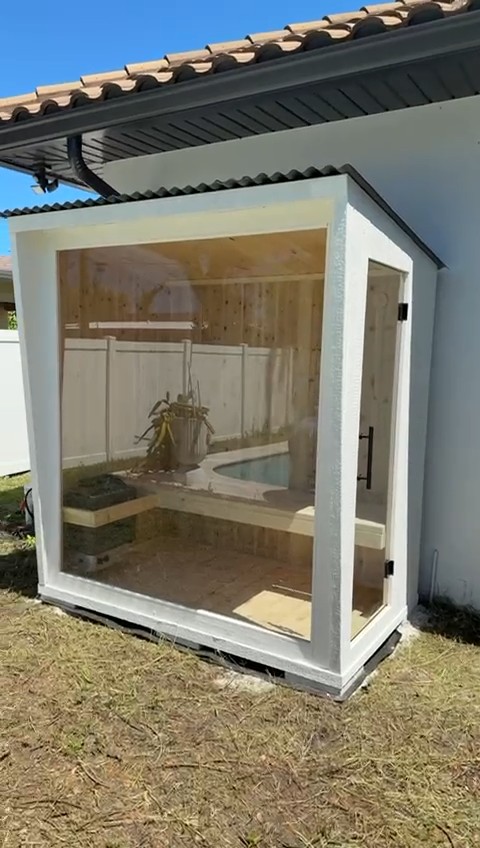 White sauna with a full-height glass front and glass side door, tucked under a house's tile-roof eave on a lawn